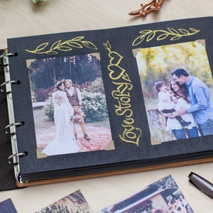 May include: Open photo album with black pages and wooden covers. The album displays two photos, one of a wedding couple and another of a family. Gold foil accents include the words "Love Story" and decorative flourishes. The album is open on a light wood surface.