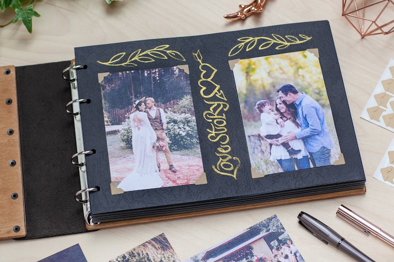 May include: Open photo album with black pages and wooden covers. The album displays two photos, one of a wedding couple and another of a family. Gold foil accents include the words "Love Story" and decorative flourishes. The album is open on a light wood surface.