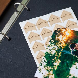 Peut inclure: Un album photo noir avec des anneaux argent&eacute;s et une feuille blanche de stickers d'angle. Les stickers sont marrons et ont un bord festonn&eacute;. Une photo d'un couple qui s'embrasse est partiellement visible sur le c&ocirc;t&eacute; droit de l'image.