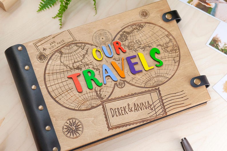 May include: A wooden photo album featuring a world map design and the words "Our Travels" spelled out in colourful letters. The album has a black leather cover with metal accents and the names "Derek & Anna" are engraved on the front.