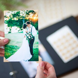 Peut inclure: Une photo de mariage d'un couple s'embrassant dans un champ vert. La photo est imprim&eacute;e sur du papier blanc avec des coins en bois et est tenue par une personne.