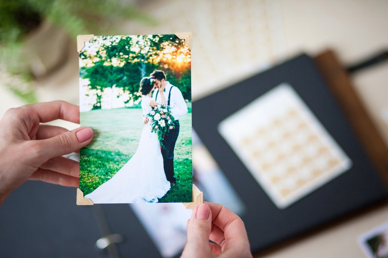 Peut inclure: Une photo de mariage d'un couple s'embrassant dans un champ vert. La photo est imprim&eacute;e sur du papier blanc avec des coins en bois et est tenue par une personne.