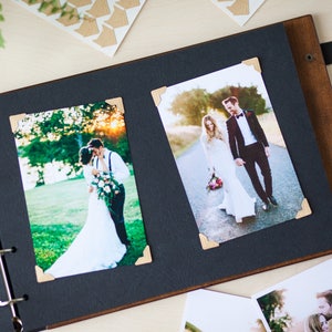 Peut inclure: Un album photo en bois brun avec des pages noires et une sangle en cuir noir. L'album est ouvert pour r&eacute;v&eacute;ler deux photos d'un couple le jour de leur mariage. Les photos sont maintenues en place par des coins dor&eacute;s.