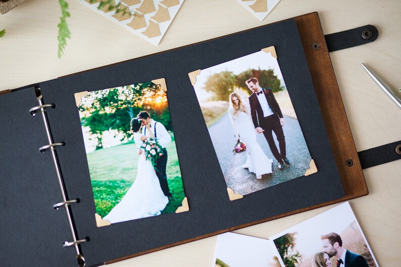 Peut inclure: Un album photo en bois brun avec des pages noires et une sangle en cuir noir. L'album est ouvert pour r&eacute;v&eacute;ler deux photos d'un couple le jour de leur mariage. Les photos sont maintenues en place par des coins dor&eacute;s.