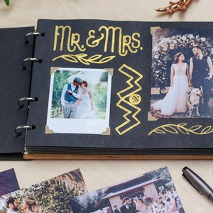 May include: A black photo album with gold lettering that says "Mr. & Mrs." The album has a wooden cover and is open to a page with two photos of a wedding couple. The photos are decorated with gold leaf accents.