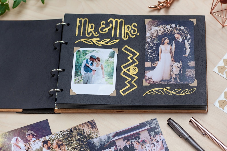 May include: A black photo album with gold lettering that says "Mr. & Mrs." The album has a wooden cover and is open to a page with two photos of a wedding couple. The photos are decorated with gold leaf accents.