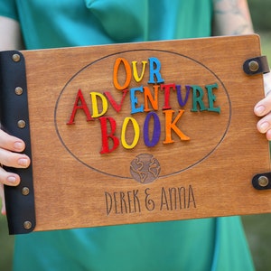 May include: Wooden adventure book with the words "OUR ADVENTURE BOOK" in colorful letters. The book has a dark brown finish, black leather binding, and the names "DEREK & ANNA" engraved at the bottom. A world map is also engraved.