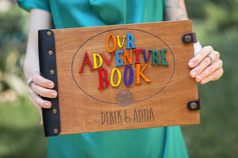 May include: Wooden adventure book with the words "OUR ADVENTURE BOOK" in colorful letters. The book has a dark brown finish, black leather binding, and the names "DEREK & ANNA" engraved at the bottom. A world map is also engraved.