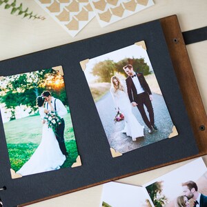 Peut inclure: Un album photo noir avec une couverture en bois et des anneaux m&eacute;talliques. L'album contient deux photos d'un couple de mari&eacute;s, l'une dans un champ et l'autre sur une route. Les photos sont d&eacute;cor&eacute;es de stickers d'angle dor&eacute;s.