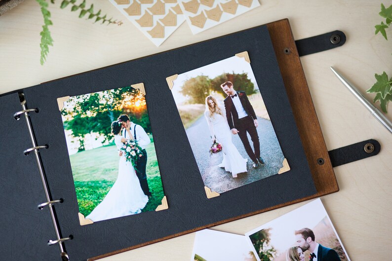 Peut inclure: Un album photo noir avec une couverture en bois et des anneaux m&eacute;talliques. L'album contient deux photos d'un couple de mari&eacute;s, l'une dans un champ et l'autre sur une route. Les photos sont d&eacute;cor&eacute;es de stickers d'angle dor&eacute;s.