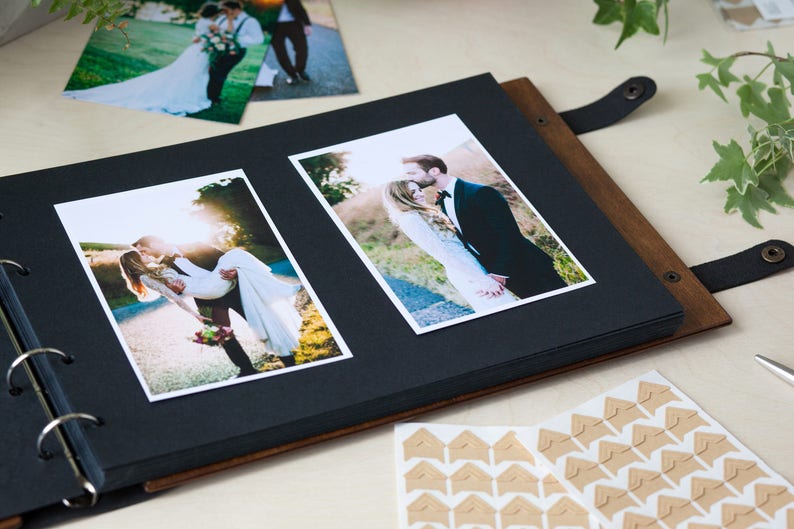 Peut inclure: Un album photo en cuir marron avec des pages noires et une reliure &agrave; anneaux. L'album est ouvert pour r&eacute;v&eacute;ler deux photos d'un couple le jour de son mariage. Les photos sont imprim&eacute;es sur du papier blanc et ont une bordure blanche.