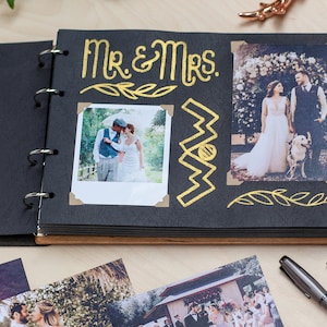 May include: Open black photo album with gold lettering that reads "Mr. & Mrs." and decorative gold accents. The album contains two wedding photos and is open on a light wood surface. Additional photos and pens are visible.