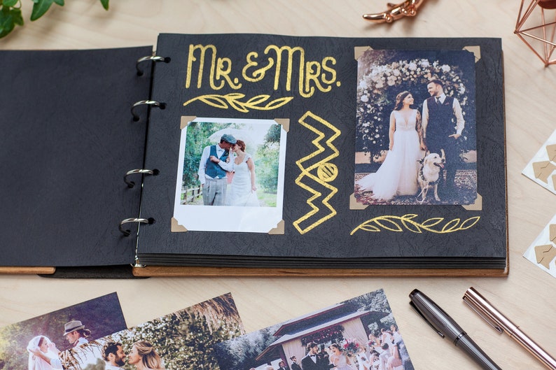 May include: Open black photo album with gold lettering that reads "Mr. & Mrs." and decorative gold accents. The album contains two wedding photos and is open on a light wood surface. Additional photos and pens are visible.
