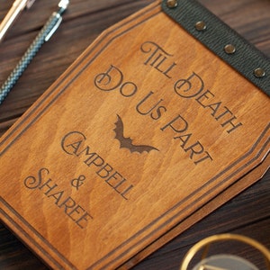 Puede incluir: Un libro de visitas de madera con forma de ata&uacute;d con una correa de cuero negro y remaches met&aacute;licos. El libro est&aacute; grabado con el texto "Till Death Do Us Part" y un gr&aacute;fico de murci&eacute;lago. Los nombres "Campbell & Sharee" tambi&eacute;n est&aacute;n grabados en el libro.