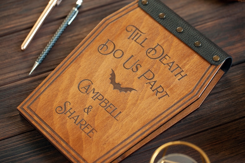 Puede incluir: Un libro de visitas de madera con forma de ata&uacute;d con una correa de cuero negro y remaches met&aacute;licos. El libro est&aacute; grabado con el texto "Till Death Do Us Part" y un gr&aacute;fico de murci&eacute;lago. Los nombres "Campbell & Sharee" tambi&eacute;n est&aacute;n grabados en el libro.