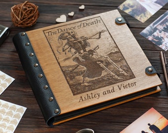 May include: A wooden photo album with a black leather cover and metal studs. The album features an engraved image of two skeletons dancing with the text "The Dance of Death" and "Ashley and Victor".