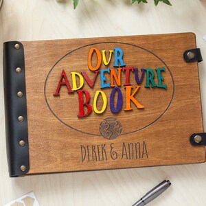 May include: Wooden photo album with the words "OUR ADVENTURE BOOK" in colorful letters. The album has a dark brown finish, black leather spine, and the names "Derek & Anna" engraved on the cover. Photos are visible in the background.