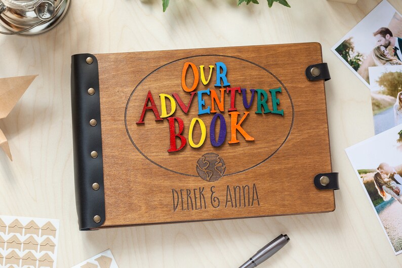 May include: Wooden photo album with the words "OUR ADVENTURE BOOK" in colorful letters. The album has a dark brown finish, black leather spine, and the names "Derek & Anna" engraved on the cover. Photos are visible in the background.