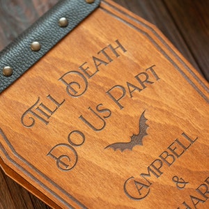 Puede incluir: Un letrero de madera con forma de ata&uacute;d con el texto "Till Death Do Us Part" y un gr&aacute;fico de murci&eacute;lago. El letrero est&aacute; personalizado con los nombres "Campbell & JaRee".