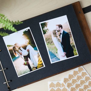 Peut inclure: Un album photo noir avec une couverture en bois et des anneaux m&eacute;talliques. L'album est ouvert pour r&eacute;v&eacute;ler deux photos d'un couple le jour de leur mariage. Les photos sont imprim&eacute;es sur du papier blanc et ont une bordure blanche. L'album est pos&eacute; sur une surface en bois.