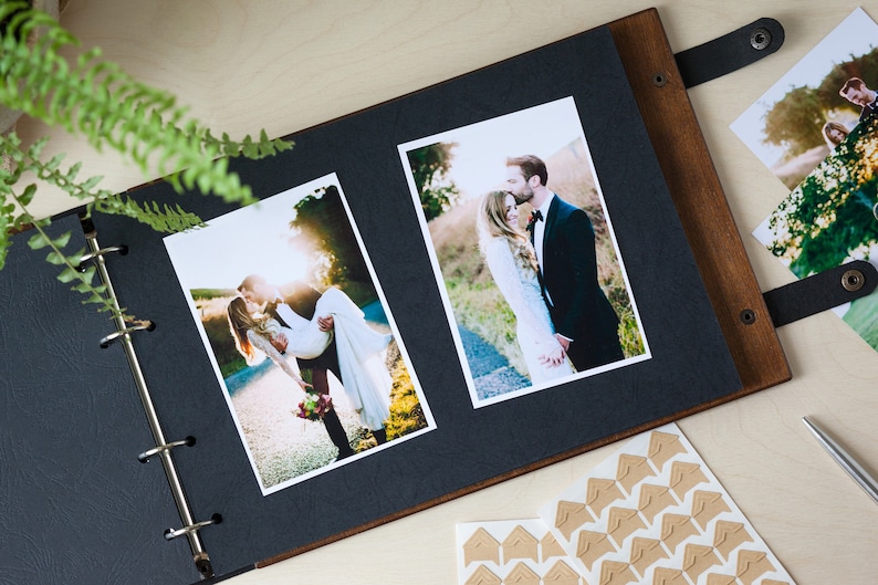 Peut inclure: Un album photo noir avec une couverture en bois et des anneaux m&eacute;talliques. L'album est ouvert pour r&eacute;v&eacute;ler deux photos d'un couple le jour de leur mariage. Les photos sont imprim&eacute;es sur du papier blanc et ont une bordure blanche. L'album est pos&eacute; sur une surface en bois.