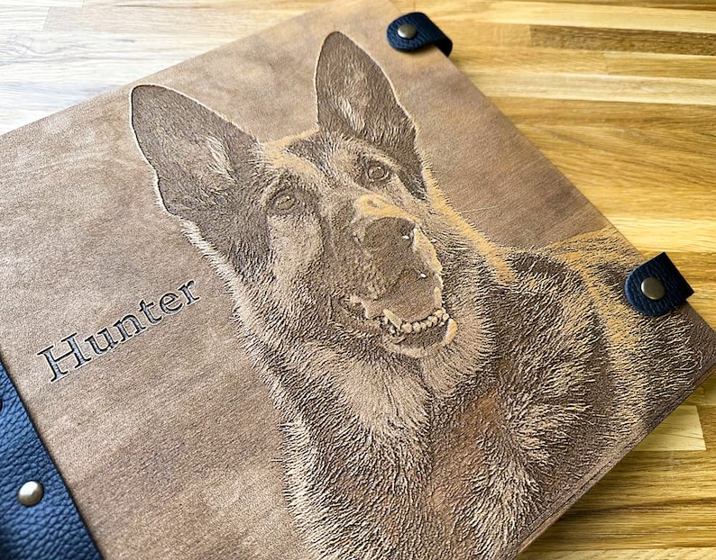 Puede incluir: Un &aacute;lbum de fotos de cuero marr&oacute;n con una correa negra y una imagen grabada con l&aacute;ser de un perro pastor alem&aacute;n con el nombre "Hunter" en la parte delantera.