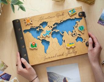 May include: A wooden photo album with a world map design and the text "Adventure Book" and "Jamaal and Shelby". The album has a black leather cover with metal rivets and features colorful icons of a plane, hot air balloon, map, compass, and a van.