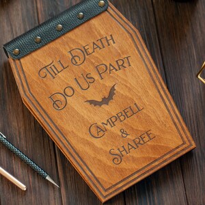 Puede incluir: Una caja de madera con forma de ata&uacute;d con una cubierta de cuero negro y tachuelas met&aacute;licas. La caja est&aacute; grabada con el texto "Till Death Do Us Part" y un s&iacute;mbolo de murci&eacute;lago. Los nombres "Campbell & Sharee" tambi&eacute;n est&aacute;n grabados en la caja.