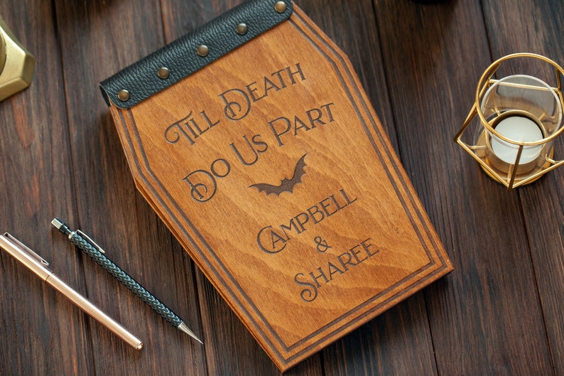 Puede incluir: Una caja de madera con forma de ata&uacute;d con una cubierta de cuero negro y tachuelas met&aacute;licas. La caja est&aacute; grabada con el texto "Till Death Do Us Part" y un s&iacute;mbolo de murci&eacute;lago. Los nombres "Campbell & Sharee" tambi&eacute;n est&aacute;n grabados en la caja.