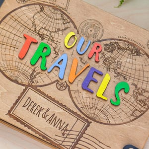 May include: A wooden photo album featuring a world map design and the words "OUR TRAVELS" spelled out in colourful letters. The album is personalised with the names "DEREK & ANNA" on a label.