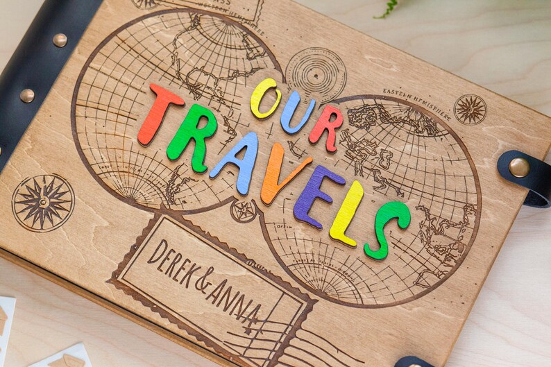 May include: A wooden photo album featuring a world map design and the words "OUR TRAVELS" spelled out in colourful letters. The album is personalised with the names "DEREK & ANNA" on a label.