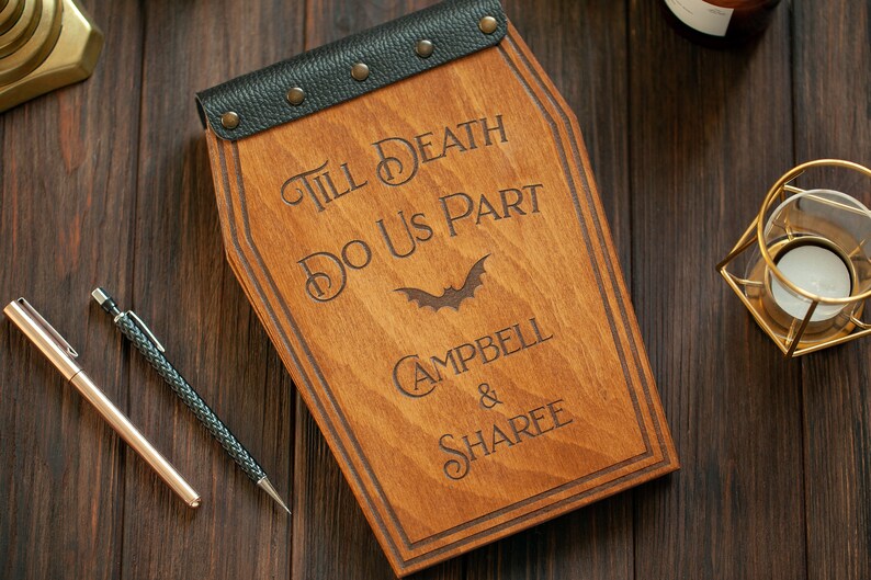 Puede incluir: Una caja de madera con forma de ata&uacute;d con una tapa de cuero negro y detalles met&aacute;licos. La caja est&aacute; grabada con el texto "Till Death Do Us Part" y los nombres "Campbell & Sharee".
