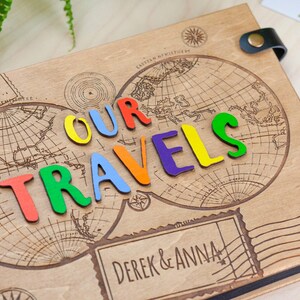 May include: A wooden photo album featuring a world map design and the words "OUR TRAVELS" spelled out in colourful letters. The album is personalised with the names "DEREK & ANNA" on the front.