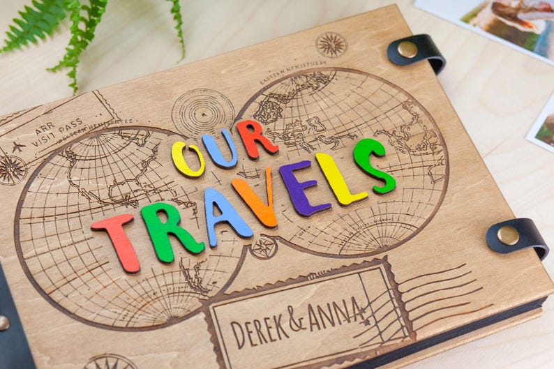 May include: A wooden photo album featuring a world map design and the words "OUR TRAVELS" spelled out in colourful letters. The album is personalised with the names "DEREK & ANNA" on the front.