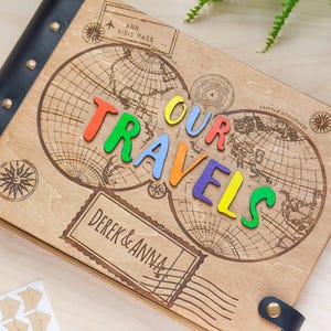 May include: A wooden photo album with a black leather cover and metal studs. The album features a world map design with the words "Our Travels" spelled out in colourful letters. The album also has a personalised inscription that reads "Derek & Anna".