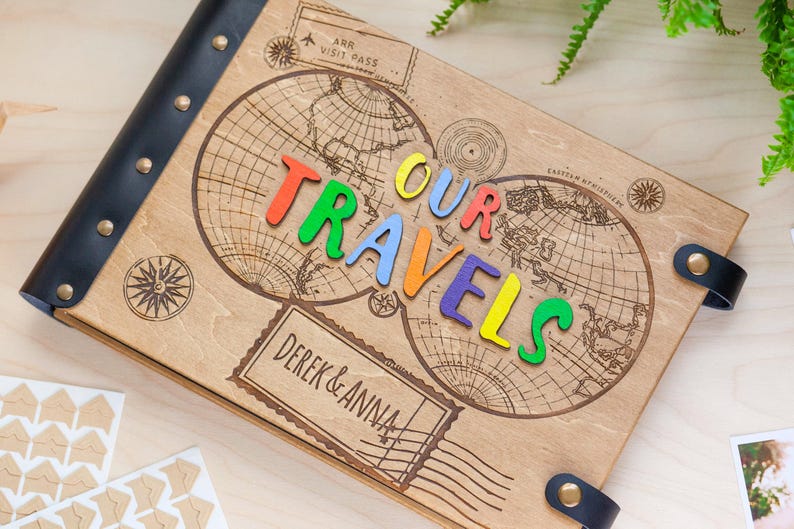 May include: A wooden photo album with a black leather cover and metal studs. The album features a world map design with the words "Our Travels" spelled out in colourful letters. The album also has a personalised inscription that reads "Derek & Anna".