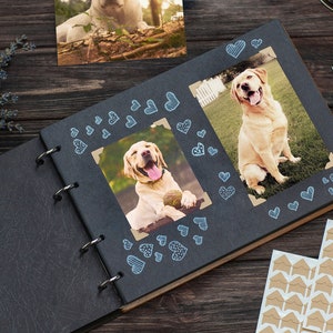 Puede incluir: Un &aacute;lbum de fotos negro con una cubierta de cuero marr&oacute;n y encuadernaci&oacute;n en espiral. El &aacute;lbum est&aacute; abierto en una p&aacute;gina con dos fotos de perros golden retriever. Las fotos est&aacute;n decoradas con pegatinas de corazones azules.