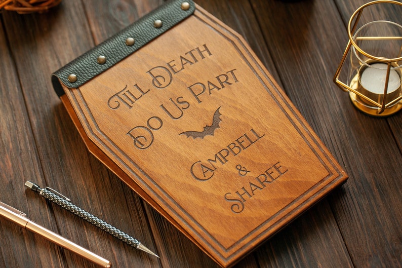 Puede incluir: Una caja de madera con forma de ata&uacute;d con una correa de cuero negro y tachuelas met&aacute;licas. La caja est&aacute; grabada con el texto "Till Death Do Us Part" y los nombres "Campbell & Sharee".