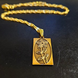 Peut inclure: Un collier en chaîne dorée avec un pendentif représentant un motif de fleur stylisé. Le pendentif est rectangulaire et a une forme légèrement ovale.