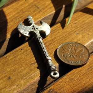 May include: A silver axe pendant with a cross design on a wooden surface next to a 1989 Canadian penny.