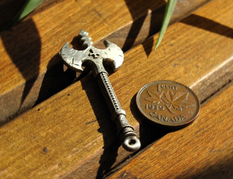 May include: A silver axe pendant with a cross design on a wooden surface next to a 1989 Canadian penny.