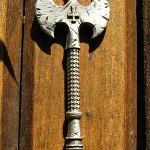 May include: A silver-colored axe with a cross on the top of the blade. The axe is mounted on a wooden surface.