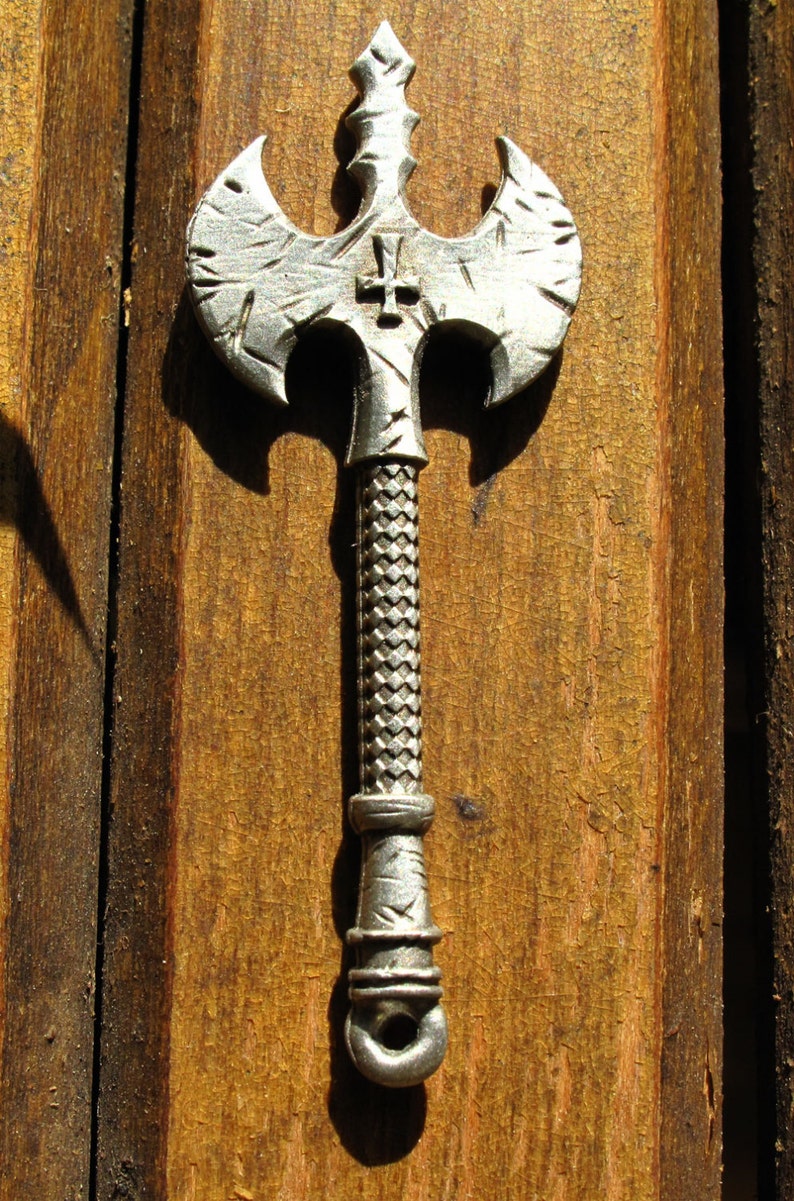 May include: A silver-colored axe with a cross on the top of the blade. The axe is mounted on a wooden surface.