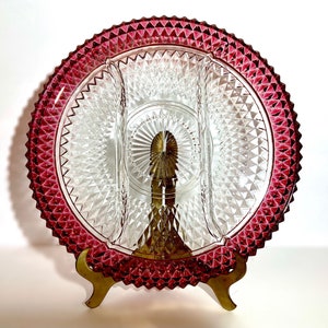 May include: A clear glass serving dish with three sections, featuring a diamond pattern. The dish has a deep red rim and is displayed on a gold metal stand.
