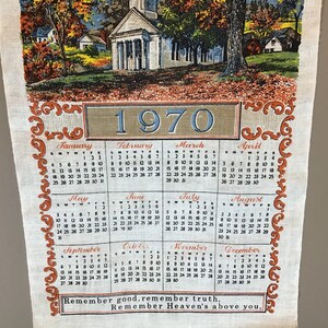 May include: A vintage 1970 calendar tea towel with a colorful illustration of a church and houses in a rural setting. The calendar features the months of the year with the days of the week and dates. The towel has a brown and orange border with a decorative design. The text "Remember good, remember truth, Remember Heaven's above you." is printed at the bottom.
