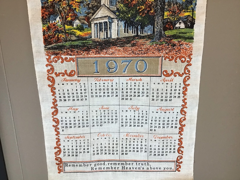 May include: A vintage 1970 calendar tea towel with a colorful illustration of a church and houses in a rural setting. The calendar features the months of the year with the days of the week and dates. The towel has a brown and orange border with a decorative design. The text "Remember good, remember truth, Remember Heaven's above you." is printed at the bottom.