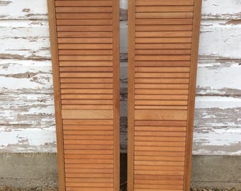 May include: Two wooden louvered shutters in a natural wood finish. The shutters are closed and are leaning against a white painted wall.