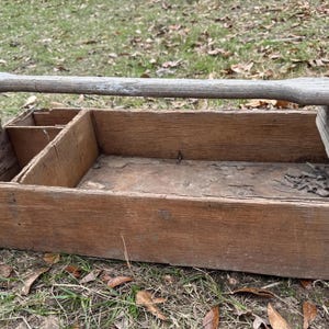 May include: A weathered wooden tool tote with a handle. The open-top box has interior dividers and is constructed from aged, light brown wood. The tote is resting on a bed of dry leaves and grass.