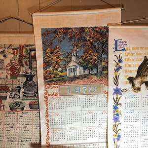 May include: Three vintage wall calendars with a white background and colorful illustrations. The top calendar features a church with a white steeple and a blue sky. The middle calendar features a black and white illustration of a blacksmith shop. The bottom calendar features a prayer with a dove and the text "Lord, make me an instrument of Thy peace."