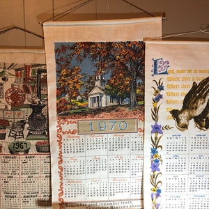 May include: Three vintage wall calendars with a country theme. The first calendar is from 1967 and features a black and white illustration of a general store. The second calendar is from 1970 and features a colorful illustration of a white church with a steeple. The third calendar features a prayer with a dove and a floral border. All three calendars have a cream background and are printed with black text.